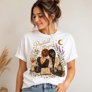 Może przedstawiać: Biały t-shirt z krótkim rękawem z grafiką przedstawiającą dwie osoby odwrócone plecami, otoczone kwiatami, świecą, kociołkiem, otwartą księgą z napisem "Trust Your Intuition" i "Magic Lies Within" oraz butelką z etykietą "Wild Elixir". Nad ilustracją znajduje się napis "Practical Magic".
