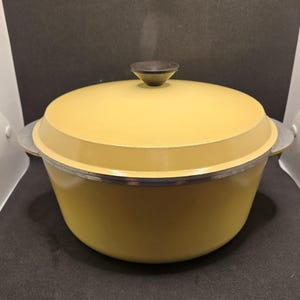 May include: A round, mustard yellow Dutch oven with a matching lid and a dark brown knob handle. The pot features two integrated side handles and a silver-toned rim where the lid meets the base, presenting a smooth, solid-colored exterior finish.