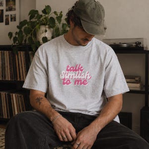 May include: A light gray heathered short-sleeve t-shirt featuring the pink text "talk Simlish to me" printed in a rounded, retro-style font across the chest. The shirt has a crew neckline and a loose, relaxed fit, made of a soft cotton blend fabric.