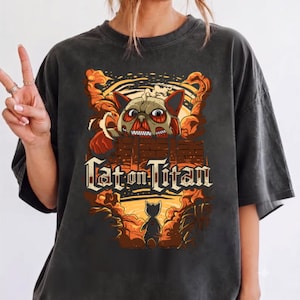 May include: A charcoal grey t-shirt featuring a graphic print of a cat with exposed muscle tissue peering over a brick wall, with the text "Cat on Titan" below. The design includes orange and yellow smoke clouds and a small cat silhouette.