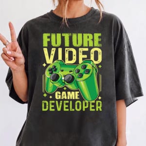 May include: A charcoal grey short-sleeve t-shirt featuring a graphic print of a bright green video game controller. The text "FUTURE VIDEO GAME DEVELOPER" is printed in bold, distressed yellow and green lettering above and below the controller.
