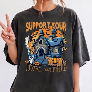May include: A charcoal gray short-sleeve t-shirt featuring a graphic print of a haunted house, pumpkins, a spider, and bats. The text reads "SUPPORT YOUR LOCAL WITCHES" in orange lettering against a large orange moon background.