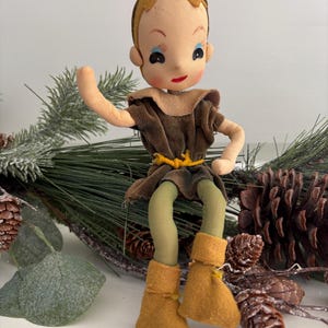 May include: A vintage cloth doll with a brown and green outfit, brown boots, and a painted face. The doll is sitting on a bed of pine branches and pine cones.