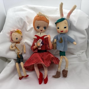 May include: Three vintage fabric dolls. A girl doll in a red polka dot dress with a white collar and red shoes. A boy doll wearing a blue sweater with a green shirt and brown boots. A bunny doll with a blue shirt and green pants.