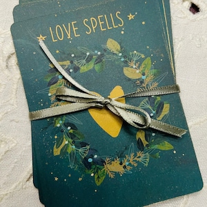 May include: A stack of cards with a green background and gold accents. The cards feature a wreath of leaves and a gold heart with the text "LOVE SPELLS" in gold lettering.