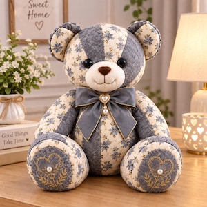 May include: A plush teddy bear constructed from patchwork fabric featuring alternating panels of cream floral print and grey textured lace. The bear has a dark brown nose, black eyes, and a grey satin bow at the neck with a heart-shaped charm.