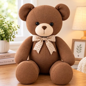 Puede incluir: Un oso de peluche marrón con un hocico beige claro y ojos redondos oscuros. El oso lleva una pajarita de cuadros beige y blancos alrededor del cuello. El juguete de peluche está hecho de una tela texturizada y está sentado en posición vertical.
