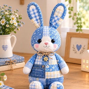 Puede incluir: Un conejo de peluche confeccionado con varios parches de tela azul y blanca, incluyendo patrones de cuadros vichy, flores y mezclilla. El conejo tiene ojos de botón negros, una nariz cosida en rosa y un pequeño dije de flor amarilla con cordel.