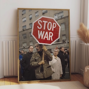 Op de afbeelding: Een ingelijste kunstdruk met een zwart-witfoto van een protestscène. Een persoon op de voorgrond houdt een groot, rood, achthoekig bord vast met de witte tekst STOP WAR. Andere personen in jassen zijn zichtbaar op de achtergrond.