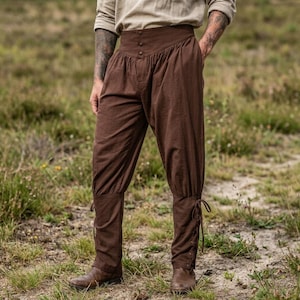 May include: Brown trousers featuring a high-waisted design with three visible buttons at the center front. The pants have a loose fit through the thighs and taper toward the lower legs, which feature adjustable lace-up closures at the ankles.