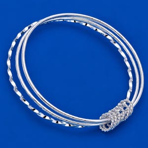 May include: A set of three interlocking silver bangles. Two of the bangles feature a smooth, polished surface, while the third bangle has a twisted, textured design. The bangles are connected by a small, circular, twisted silver ring component.
