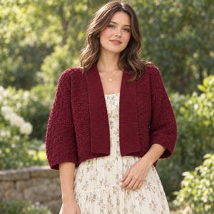 Op de afbeelding: Een diepbordeauxrood gebreid vest met een gestructureerd breipatroon, een open voorzijde en driekwartmouwen. Het kledingstuk wordt gedragen over een crèmekleurige jurk met een fijne bloemenprint en een rok met verschillende lagen.