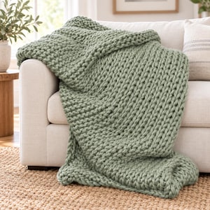 Op de afbeelding: Een saliegroene, grof gebreide plaid met een dikke, getextureerde garenstructuur. De deken heeft een opvallend, oversized lussteekpatroon en is gedrapeerd over de armleuning en zitting van een lichtgekleurde gestoffeerde bank.