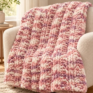 May include: A chunky knit throw blanket featuring a variegated color pattern of soft pink, cream, and lavender yarn. The blanket is constructed with thick, plush loops of yarn arranged in vertical rows, creating a textured and soft appearance.