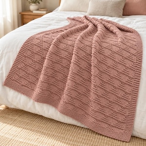 Könnte beinhalten: Eine altrosa gestrickte Wohndecke mit einem sich wiederholenden geometrischen Dreiecksmuster aus strukturierten Strickmaschen. Die Decke hat einen breiten, glatt gestrickten Rand und liegt in weichen Falten über einer weißen Tagesdecke drapiert.