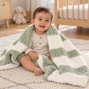 Puede incluir: Una manta para bebé suave y texturizada con anchas rayas horizontales en verde salvia y crema. La manta tiene una construcción de hilo grueso y rizado, y está colocada sobre un bebé sonriente que viste una camisa de botones color crema.
