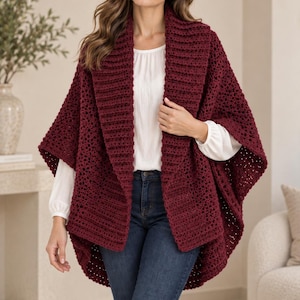 May include: A deep burgundy crochet wrap featuring a wide, ribbed collar and an open-front design. The garment is layered over a white long-sleeve blouse and dark blue denim jeans, showcasing a textured, open-stitch pattern throughout the body.