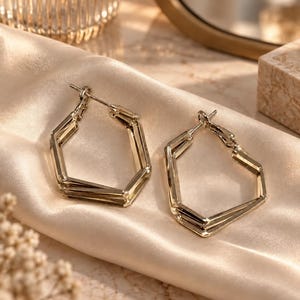 May include: A pair of gold-toned hoop earrings featuring a geometric hexagonal shape. Each earring is constructed from multiple layered metal bands that form a sharp, angular silhouette. The earrings include a standard post and hinged closure mechanism.