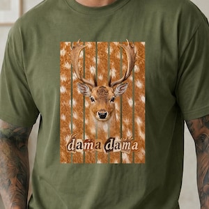 May include: An olive green short-sleeve t-shirt featuring a central graphic of a fallow deer head centered over a vertical striped background with a spotted pattern. The text "dama dama" is printed in a brown serif font below the deer's head.