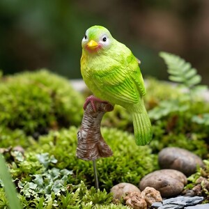 May include: A small, bright lime green bird figurine with a yellow beak and white-rimmed eyes, perched atop a textured brown branch-shaped stake. The bird features detailed feather patterns and pinkish feet, designed to be inserted into the ground.