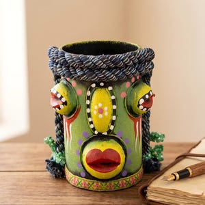 May include: A cylindrical green desk organizer featuring a stylized face with a prominent red mouth, yellow oval accents, and two protruding side features. The top rim is wrapped in thick, dark blue and white twisted rope trim.