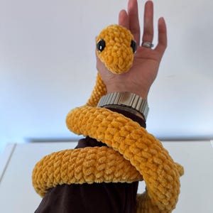 May include: A long, plush, mustard yellow snake toy crafted from soft, textured yarn. The snake features a rounded head with two large, glossy black plastic eyes and a long, coiled body that wraps around the arm, showcasing a consistent knit pattern.