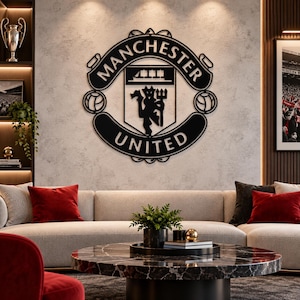 May include: A black metal wall art piece featuring the Manchester United football club crest. The circular design includes the text Manchester United, a central shield with a ship icon, and a devil figure holding a trident, flanked by two footballs.