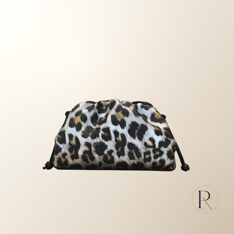 May include: A small, structured pouch featuring a prominent black, white, and gold leopard print pattern. The bag has a gathered, rounded top silhouette with black drawstring cords on both sides, creating a cinched effect at the closure.