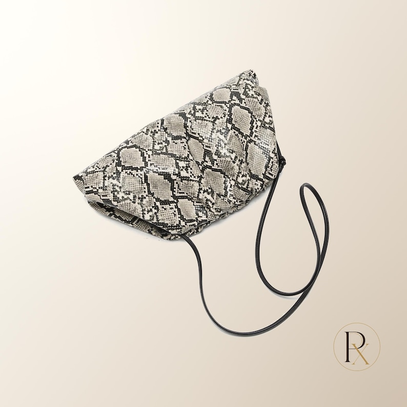 May include: A soft, gathered clutch bag featuring a grey, black, and white snakeskin pattern. The bag has a rounded, voluminous shape and includes a thin, black cord shoulder strap that hangs down from the side of the main body of the bag.
