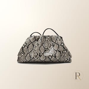 May include: A structured clutch bag featuring a grey, black, and white snakeskin pattern. The bag has a gathered, pouch-like silhouette with a hinged frame closure and a thin, black shoulder strap attached to the top. The design is textured.