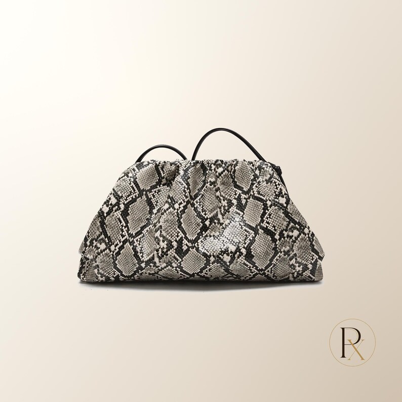 May include: A structured clutch bag featuring a grey, black, and white snakeskin pattern. The bag has a gathered, pouch-like silhouette with a hinged frame closure and a thin, black shoulder strap attached to the top. The design is textured.