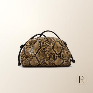 May include: A pouch-style handbag featuring a tan and black snakeskin pattern. The bag has a gathered, rounded top closure and two thin black cord handles that extend from the sides, each finished with a small knot at the base of the bag.