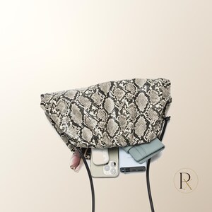 May include: A slouchy, soft-structured clutch bag featuring a grey and black snakeskin print pattern. The open interior reveals a light-colored smartphone, a pair of white wireless earbuds in a case, a slim light blue wallet, and a black power bank.