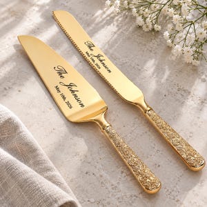 May include: A gold-toned cake knife and server set with ornate, textured handles featuring floral detailing. Both pieces are engraved with the text "The Johnson" and "May 10th, 2026" in a black script font on the smooth, polished metal blades.