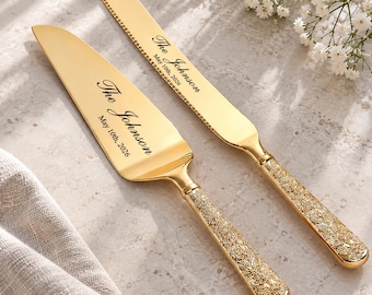 Könnte beinhalten: Ein goldenes Tortenmesser- und Tortenheber-Set mit verzierten, strukturierten Griffen mit Blumenmuster. Beide Teile sind mit dem Text "The Johnson" und "May 10th, 2026" in schwarzer Schreibschrift auf den glatten, polierten Metallklingen graviert.