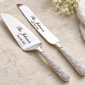 Puede incluir: Un juego de cuchillo y pala para pastel en plata con mangos ornamentados con relieves florales. Las hojas están grabadas con el texto The Johnson y la fecha May 10th, 2026. El cuchillo tiene un borde dentado y la pala una hoja triangular.