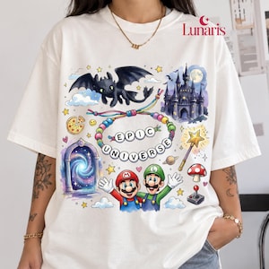 Puede incluir: Una camiseta blanca de manga corta con un estampado gráfico colorido que muestra un dragón negro, un castillo, un portal, una varita mágica, un hongo, un joystick, una porción de pizza y dos personajes con el texto EPIC UNIVERSE.