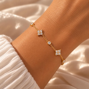 Può includere: Un bracciale a catena dorata con pietre trasparenti a taglio rotondo che si alternano tra gruppi a forma di fiore a quattro petali e singole pietre incastonate. Sottili maglie metalliche collegano ogni elemento decorativo in un motivo.