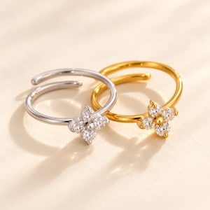 Può includere: Due anelli aperti, uno color argento e uno color oro, ciascuno caratterizzato da un motivo a fiore a quattro petali al centro. Il design floreale su entrambi gli anelli è composto da quattro pietre chiare a taglio rotondo attorno a una perla.