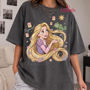 Könnte beinhalten: Ein anthrazitfarbenes T-Shirt mit einem Grafikdruck von Rapunzel mit langem blonden Haar, einem grünen Chamäleon und schwebenden Laternen. Der Text "Rapunzel" ist links in Schreibschrift gedruckt, der Markenname "Lunaris" steht oben rechts.