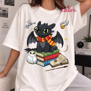 Può includere: Una t-shirt color bianco sporco con la grafica di un drago nero che indossa occhiali tondi e una sciarpa a righe rosse e gialle, seduto su una pila di libri verdi, rossi e blu. Sono presenti un gufo bianco, una chiave volante, un boccino d'oro e una bacchetta.