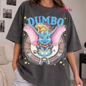 Puede incluir: Una camiseta de manga corta gris carbón con un gráfico del personaje Dumbo con grandes orejas rosas, un sombrero amarillo y un cuello rojo. El diseño incluye el texto Dumbo, un fondo de carpa de circo y estrellas amarillas al frente.