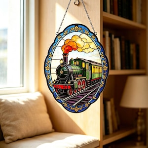Könnte beinhalten: Ein ovaler Buntglas-Sonnenfänger mit einer grünen und gelben Dampflokomotive auf Bahngleisen. Der Zug stößt orangefarbenen und gelben Rauch aus. Das Stück ist von einem blauen Rand mit goldenen Blumenakzenten umrahmt und hängt an einer schwarzen Kette.
