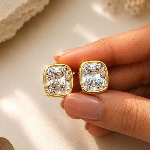 May include: A pair of stud earrings featuring cushion-cut clear stones set in gold-toned metal bezels. The earrings have a smooth, polished finish and a square shape with rounded corners, reflecting light from the faceted surfaces of the stones.