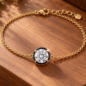 Könnte beinhalten: Ein goldenes Kettenarmband mit einem einzelnen runden, klaren, facettierten Stein, der in einer schwarzen kreisförmigen Fassung sitzt. Das Armband besteht aus einer feinen Gliederkette, einem Karabinerverschluss und einer Verlängerungskette mit einem kleinen runden Goldanhänger.