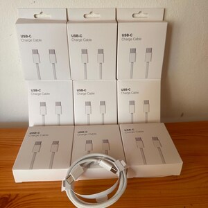 Peut inclure: Huit boîtes rectangulaires blanches étiquetées USB-C Charge Cable, chacune présentant un graphique d'un câble blanc avec deux connecteurs USB-C. Un seul câble de charge USB-C tressé blanc est enroulé devant les boîtes sur une surface en bois.