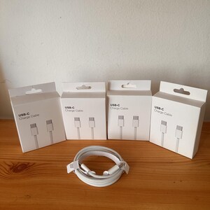 Peut inclure: Quatre boîtes rectangulaires blanches étiquetées USB-C Charge Cable, chacune illustrant un câble USB-C blanc. Devant les boîtes se trouve un câble USB-C blanc enroulé avec une texture tressée et des connecteurs aux deux extrémités.
