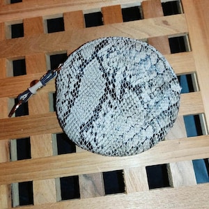 Puede incluir: Un monedero circular con un patrón de piel de serpiente en gris, negro y blanco. La bolsa incluye un pequeño tirador de cremallera metálico unido a una correa corta y oscura con un cierre metálico, lo que permite cerrar el accesorio redondo.
