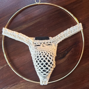 Op de afbeelding: Een crèmekleurige gehaakte string met een opengewerkt netpatroon, opgehangen in een cirkelvormige goudkleurige metalen ring. Het kledingstuk is gemaakt van garen en vertoont een gestructureerd, ingewikkeld steekpatroon over het voorpaneel.