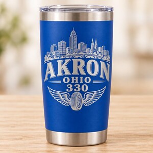 May include: A blue stainless steel tumbler with a clear lid, featuring a white laser-etched design of the Akron, Ohio skyline, the text "AKRON OHIO 330", and a graphic of a tire with wings centered below the text on the front of the vessel.